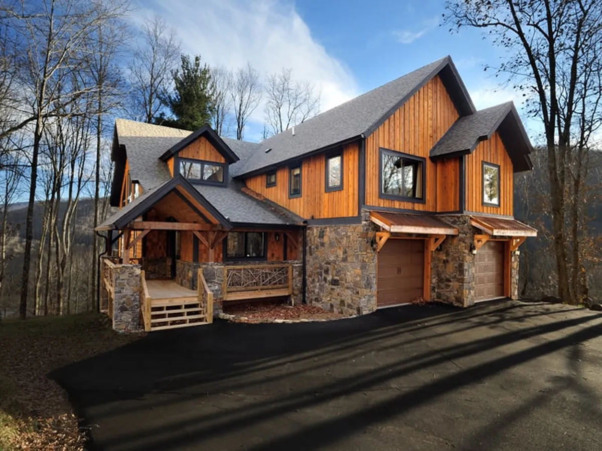 The hero image shows the inviting exterior of a custom timber-framed townhome, featuring a warm wooden facade paired with elegant stone accents, expansive windows, and a beautifully landscaped entrance surrounded by mountain scenery. The setting suggests a welcoming space ideal for gatherings and relaxation, with porches perfect for taking in the sunrise or enjoying evenings under the stars.