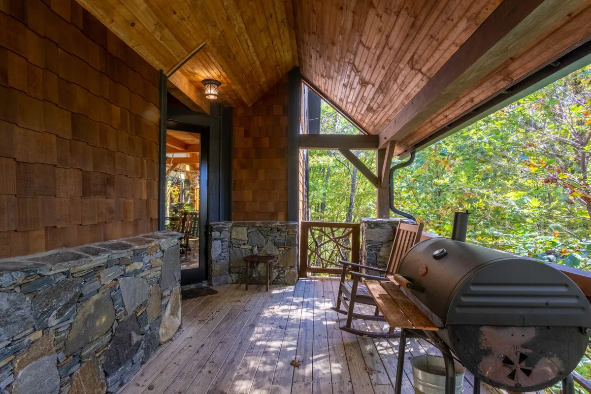A peaceful porch scene featuring rocking chairs, a grill for barbecues, a stone wall, and sprawling views of the surrounding nature. The setting exudes calm and is perfect for both relaxing and social gatherings with friends or family.