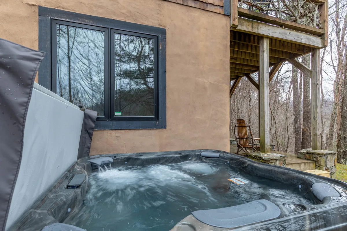 A serene outdoor scene featuring a private wooden deck nestled among the trees, with a luxurious hot tub as the focal point. Stone steps lead up to the deck, and the surrounding forest creates a peaceful, secluded atmosphere ideal for relaxation and enjoying nature.