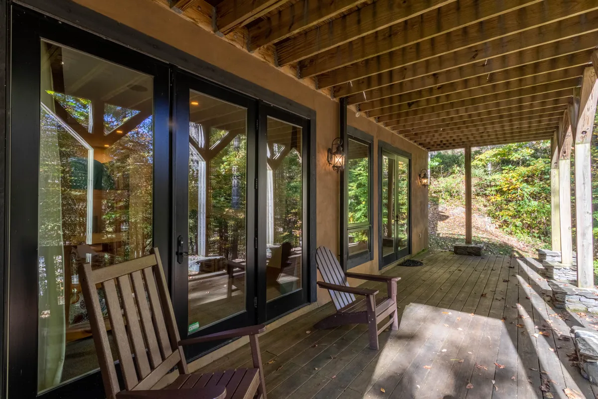 A tranquil outdoor scene featuring wooden rocking chairs on a spacious deck, with large glass doors opening to the natural surroundings. The setting is bathed in natural light, offering views of greenery and inviting guests to relax outside.