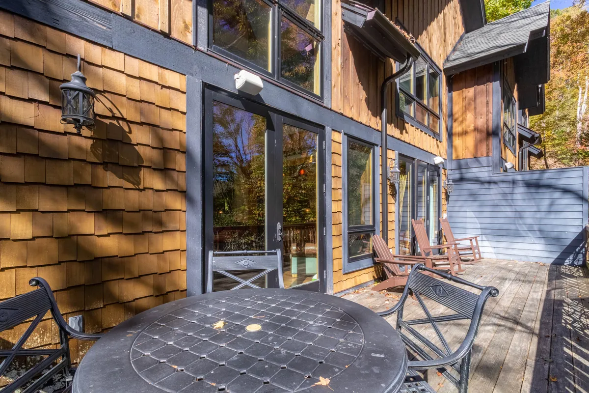 A welcoming exterior setting with a wooden deck furnished with a table and chairs, large windows connecting the indoor and outdoor spaces, and lush natural surroundings. The scene invites leisurely outdoor dining or quiet moments enjoying the mountain air.
