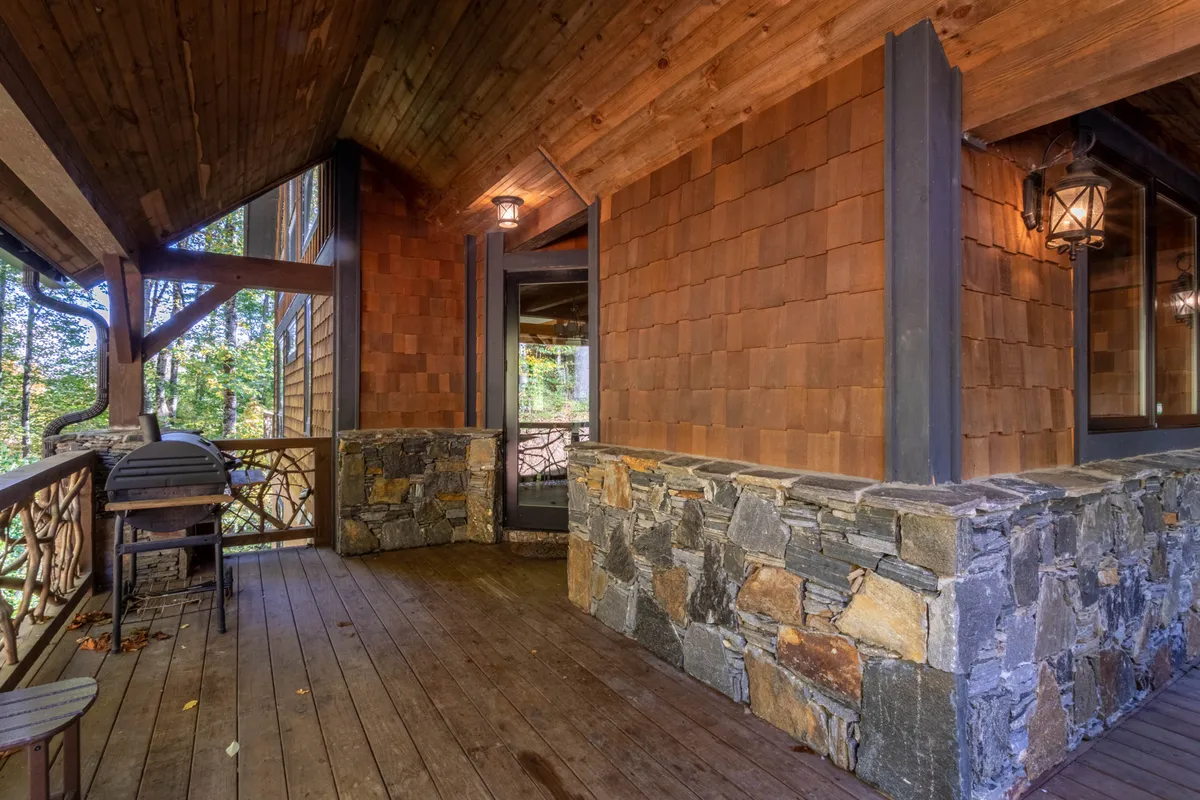 A cozy, inviting wooden deck with a sturdy stone wall and shingle siding. Soft lantern light glows at twilight, the deck surrounded by trees, accessible through a glass door. The atmosphere promises comfort and a connection to nature.