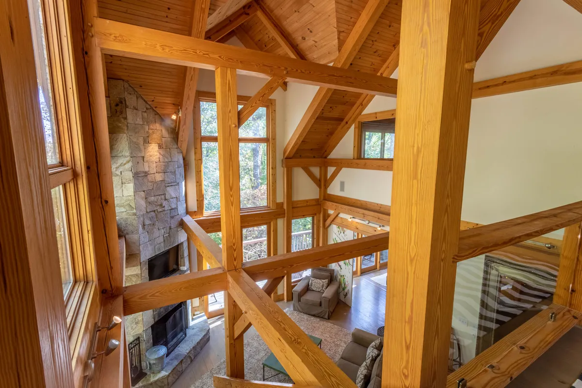 A spacious Great Room with high wooden beams, a large stone fireplace, expansive windows letting in natural light, and a comfortable seating area arranged for conversation and relaxation. The setting is warm, inviting, and perfect for gatherings, with mountain views visible through the windows.