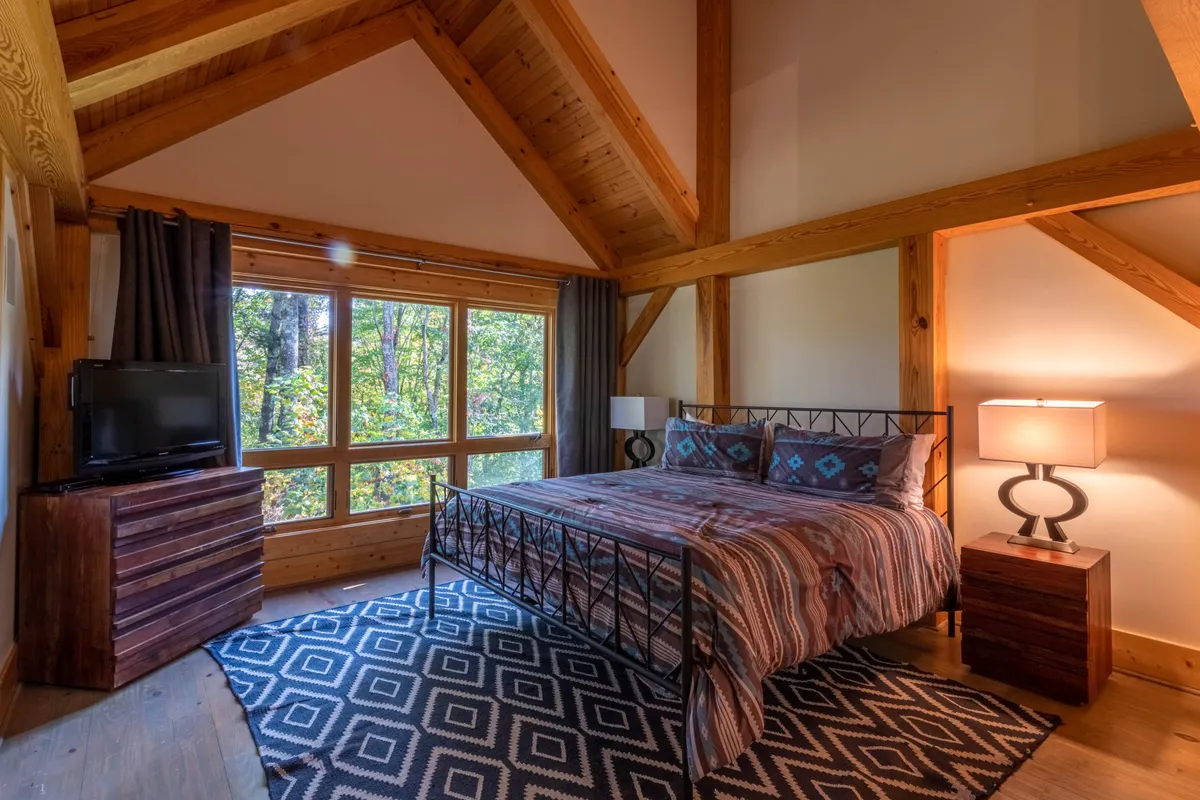 A master suite featuring a large bed with patterned bedding, wooden accents, and expansive windows that look out onto a serene forest view. A cozy nightstand with a lamp sits beside the bed, and the whole space is bathed in warm, inviting light—ideal for relaxing mornings with coffee or evenings unwinding after adventure.