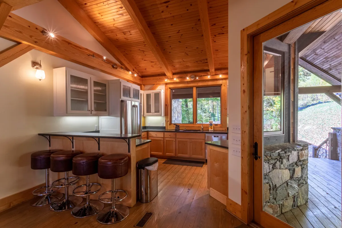 A view of the kitchen area featuring a large island with bar stools, wooden beams overhead, a stone accent wall, and large windows that fill the room with light. The space is designed for group cooking, entertaining, and casual dining.