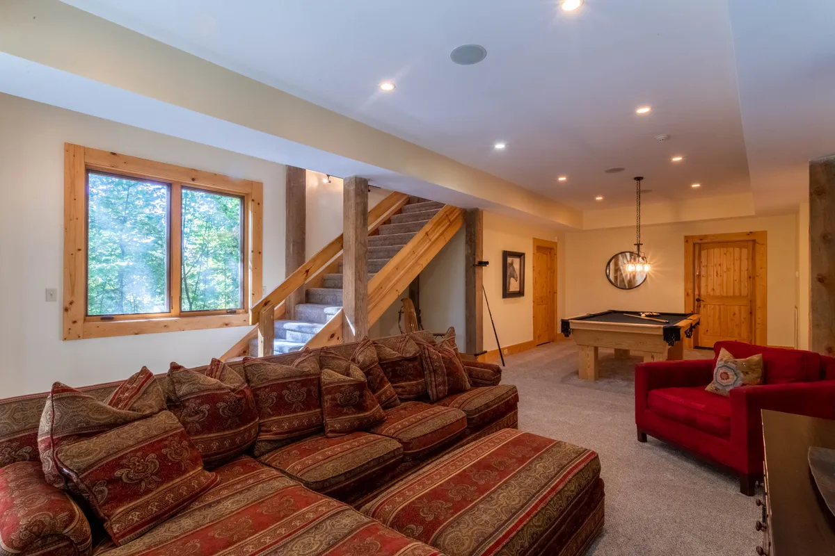 This inviting entertainment den features a large sectional sofa, a pool table, a staircase leading upstairs, a red accent chair, and decorative wall art. The space is designed for relaxation and fun, offering plenty of room for group activities, movie nights, and friendly competitions.