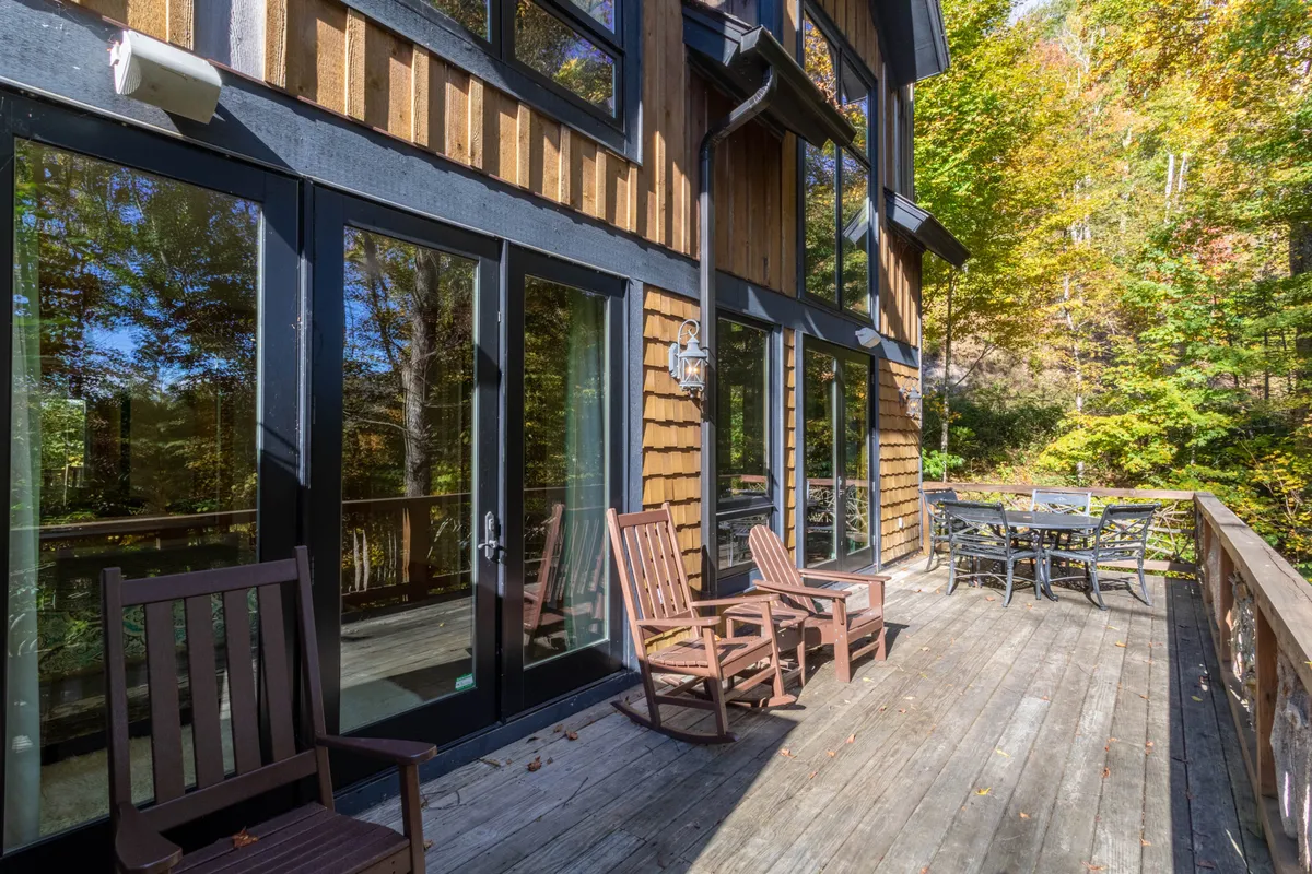 An exterior deck featuring wooden rocking chairs, a dining table, large windows reflecting the interior glow, and surrounding trees. The space is ideal for outdoor dining, relaxing with morning coffee, or watching the sunset over the Tennessee Valley.