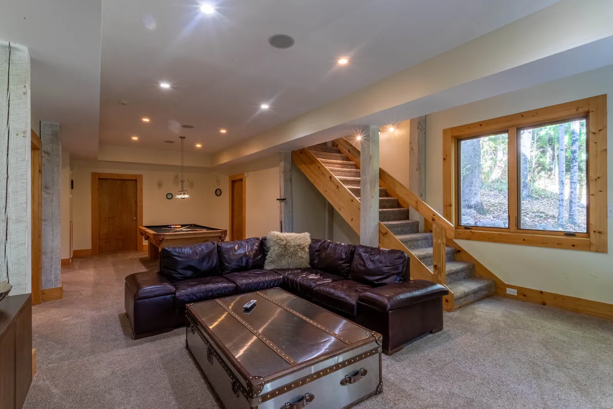 A cozy den with a large leather sectional sofa, a prominent pool table at center, a trunk coffee table, and a large window offering forest views. The arrangement encourages both playing pool and relaxing with friends or family.